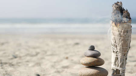 Dried white sage smoke, smudge stick burning in soft focus with bokeh, aroma smudging close up. Rock balancing on ocean beach, stones stacking by sea water waves. Zen pyramid of pebbles on sandy shoreの写真素材