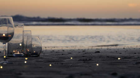 Candle flame lights in glass, romantic beach date by California ocean waves, summer sea water. Candlelight looped cinemagraph. Wineglass, glass for wine on sand. Cozy lounge garland, sunset.の写真素材