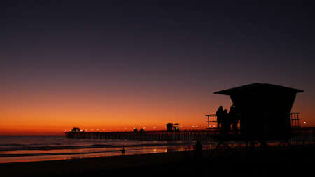 Young teen girls silhouettes near lifeguard tower, friends on pacific ocean beach, sunset dusk in Oceanside, California USA. Unrecognizable teenagers, people and twilight gradient purple violet sky.の写真素材