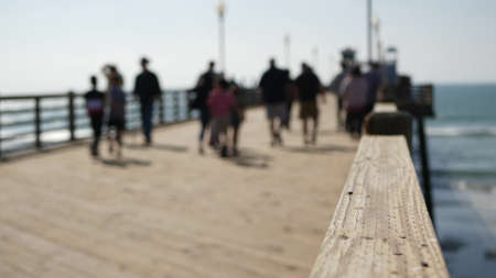 Railings of old wooden pier, people walking on waterfront boardwalk, Oceanside beach atmosphere, California coast USA. Defocused seascape, pacificocean water waves. Los Angeles summertime vacations.の写真素材