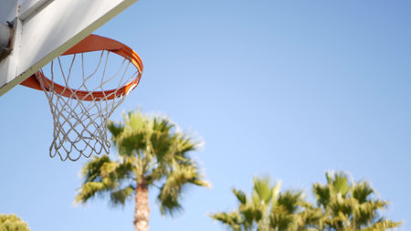 Basketball court outdoors, orange hoop, net and backboard for basket ball game outside. Recreational sport equipment on streetball field or playground. Blue sky and beach palm trees, California USA.の写真素材