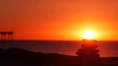 Stack of pebble stones, sandy ocean beach, sunset sky. Rock balancing in sun light, sea water waves....
