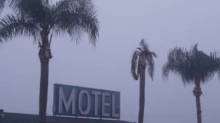 Retro neon sign of motel or hotel on road, foggy misty weather in California, USA. Palm trees in...