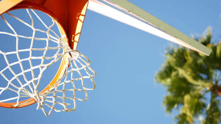 Basketball court outdoors, orange hoop, net and backboard for basket ball game outside. Recreational sport equipment on streetball field or playground. Blue sky and beach palm trees, California USA.の写真素材