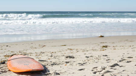 Surfboard for surfing lying on beach sand, California coast, USA. Ocean waves and orange surf board...