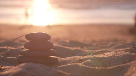 Stack of pebble stones, sandy ocean beach, sunset sky. Rock balancing in sun light, sea water waves. Stones staking in pyramid pile. Zen meditation and harmony in balance. Calm tranquil atmosphere.の写真素材