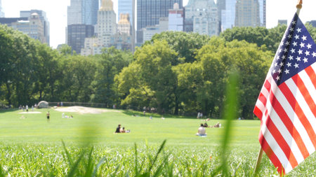 American flag on green grass in Central Park, New York, USA. Sheep Meadow, high-rise buildings and people. Skyline cityscape, Manhattan Midtown skyscrapers. United States symbol and business district.の写真素材