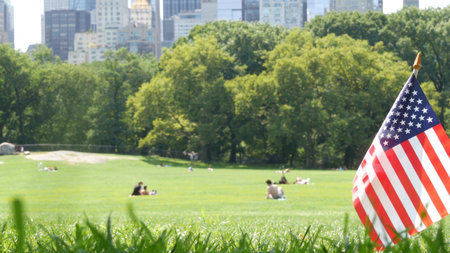 American flag on green grass in Central Park, New York, USA. Sheep Meadow, high-rise buildings and people. Skyline cityscape, Manhattan Midtown skyscrapers. United States symbol and business district.の写真素材