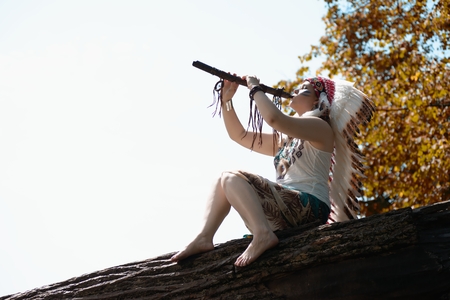Young woman in war bonnet headdress of American Indian plays on a tree a fluteの写真素材