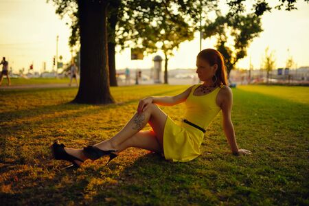 The beautiful girl in a yellow dress sits on a grass in beams of the sunset sunの写真素材