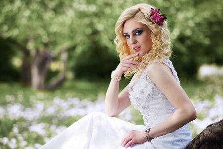 Beautiful young bride in white dress sits on tree in summer green parkの写真素材