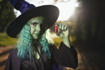 Young girl with green hair and skin suit of witch in forest holds small bottle with red potion. Halloween timeの写真素材