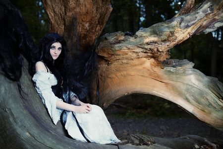 Young beautiful witch with long dark hair in root of old treeの写真素材