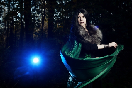 Young girl looks as witch and blows raincoat in dark forestの写真素材