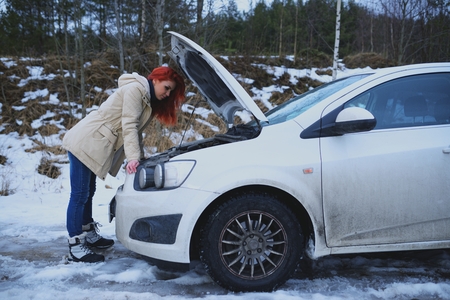 Young redhead girl looks under cowl of broken car on rural roadの写真素材