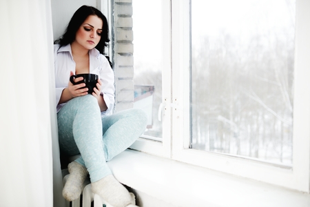 Young woman sits on window sill with cup in hands and looks out of window. Morning of winter.の写真素材