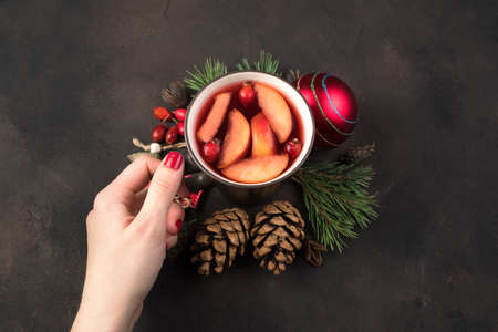 Christmas composition, a womans hand puts a mug in the center of Christmas toys and pine branches on a brown concrete background. The concept of New year and Christmas.の写真素材