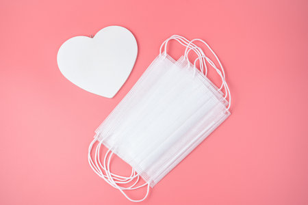 White heart and a protective mask on the pink background. Top view, romantic background. Concept...