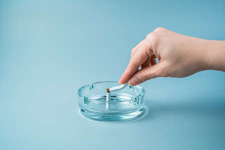 A hand breaks a cigarette in an ashtray on a blue background. Side view with copy space. The concept...