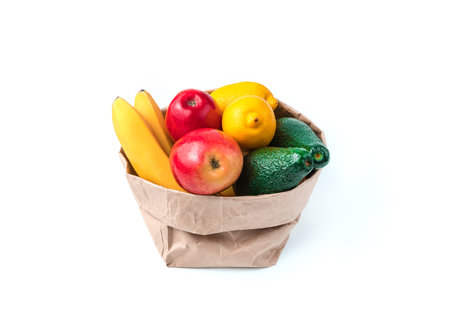 The craft bag is filled with fruits: bananas, avocados, lemons and apples on a white background. Side view. The concept of food products.の写真素材