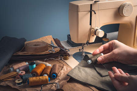 Tailors hands, thread, fabric and sewing machine on a blue background. Side view with copy space.