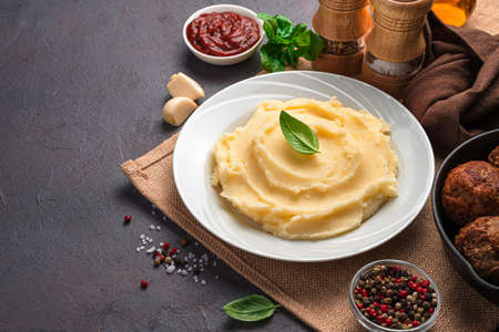 Mashed potatoes with fresh basil in a white plate on a dark background with sauce, spices and garlic. Side view, copy space.の写真素材