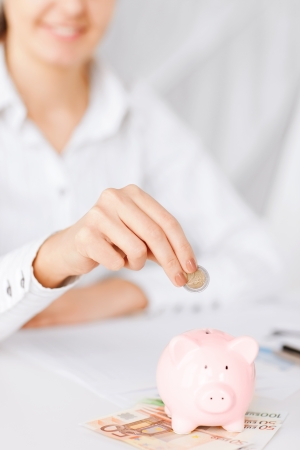 business, office, household, school, tax and education concept - woman hand putting coin into small piggy bankの写真素材