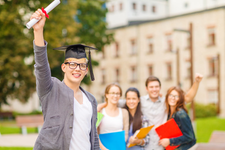 education, campus and teenage concept - smiling teenage boy in corner-cap and eyeglasses with diploma and classmates on the backの写真素材