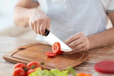 cooking and home concept - close up of male hand cutting tomato on cutting board with sharp knife