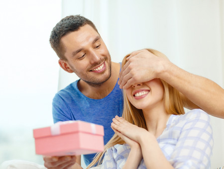 love, holiday, celebration and family concept - smiling man surprises his girlfriend with present at homeの写真素材