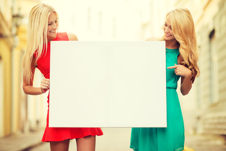summer holidays, , travel, tourism and advertisement concept - two happy blonde women with blank white board in the cityの写真素材