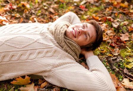 season, happiness and people concept - close up of smiling young man lying on ground or grass and fallen leaves in autumn parkの写真素材