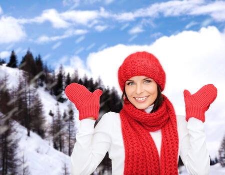 happiness, winter holidays, tourism, travel and people concept - smiling young woman in red hat and mittens over snowy mountains backgroundの写真素材