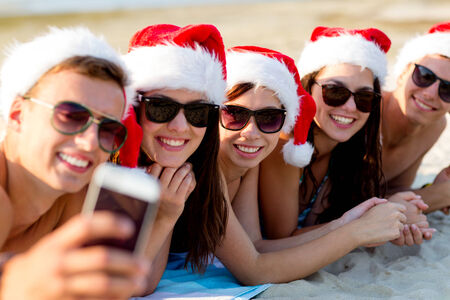 friendship, summer vacation, holidays, technology and people concept - group of friends in santa helper hats taking selfie with smartphone on beachの写真素材