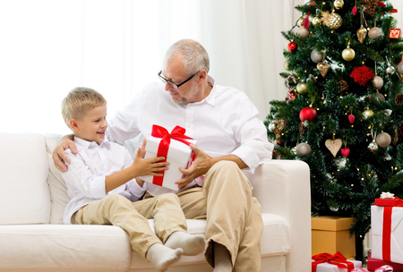 family, holidays, generation, christmas and people concept - smiling grandfather and grandson with gift box sitting on couch at homeの写真素材