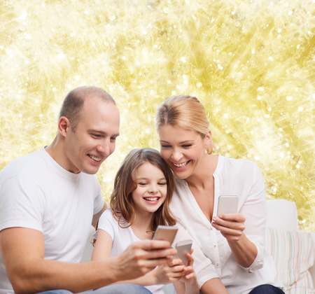 family, holidays, technology and people concept - smiling mother, father and little girl with smartphones over yellow lights backgroundの写真素材