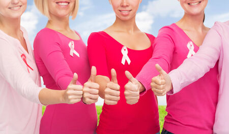 healthcare, people, gesture and medicine concept - close up of smiling women in blank shirts with pink breast cancer awareness ribbons showing thumbs up over blue sky and grass backgroundの写真素材