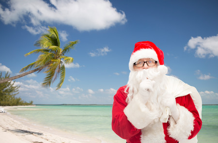 christmas, holidays, travel and people concept - man in costume of santa claus with bag making hush gesture over tropical beach backgroundの写真素材