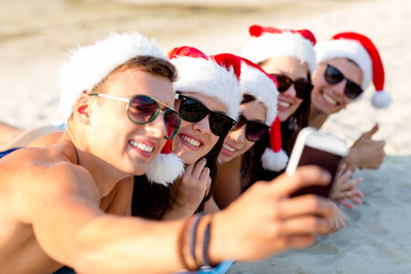 friendship, summer vacation, holidays, technology and people concept - group of friends in santa helper hats taking selfie with smartphone on beachの写真素材
