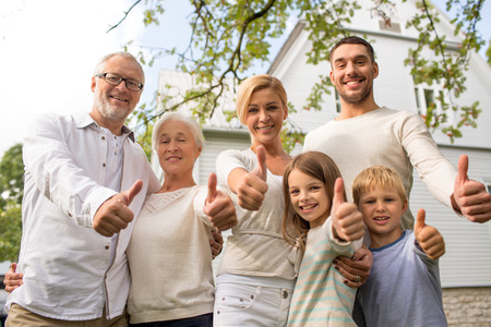 family, happiness, generation, home and people concept - happy family standing in front of house and showing thumbs up outdoorsの写真素材