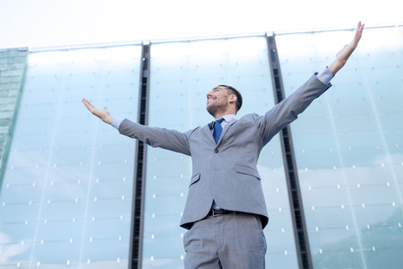 business, people and education concept - young smiling businessman with raised hands over office buildingの写真素材