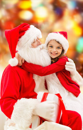 holidays, christmas, childhood and people concept - smiling little girl hugging with santa claus over red lights backgroundの写真素材