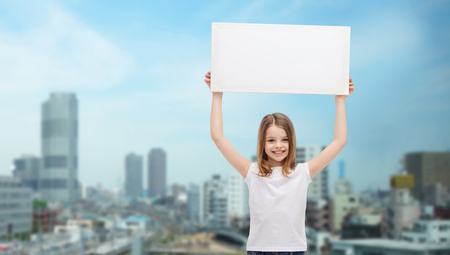 advertisement, childhood, happiness and people concept - smiling little child in white t-shirt holding blank board over city backgroundの写真素材
