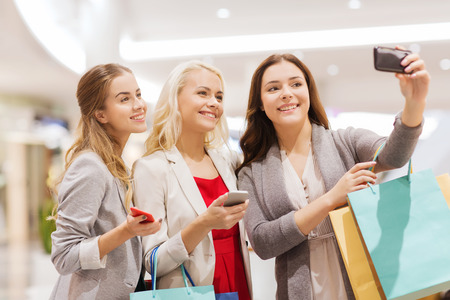 sale, consumerism, technology and people concept - happy young women with smartphones and shopping bags taking selfie in mallの写真素材
