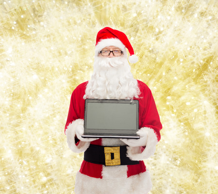 christmas, advertisement, technology, and people concept - man in costume of santa claus with laptop computer over yellow lights backgroundの写真素材