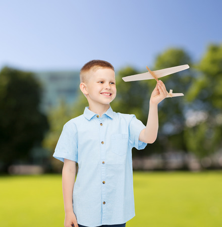 dreams, future, hobby, nature and childhood concept - smiling little boy holding wooden airplane model in his hand over park backgroundの写真素材