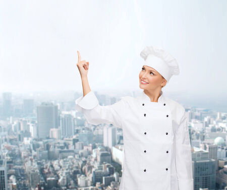 cooking, advertisement and people concept - smiling female chef, cook or baker pointing finger up to something over city backgroundの写真素材