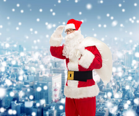 christmas, holidays and people concept - man in costume of santa claus with bag looking far away over snowy city backgroundの写真素材