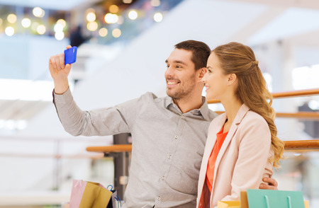 sale, consumerism, technology and people concept - happy young couple with shopping bags and smartphone taking selfie in mallの写真素材