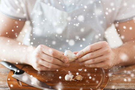 cooking, food, people and home concept - close up of male hands taking off peel of garlic on cutting board in kitchenの写真素材